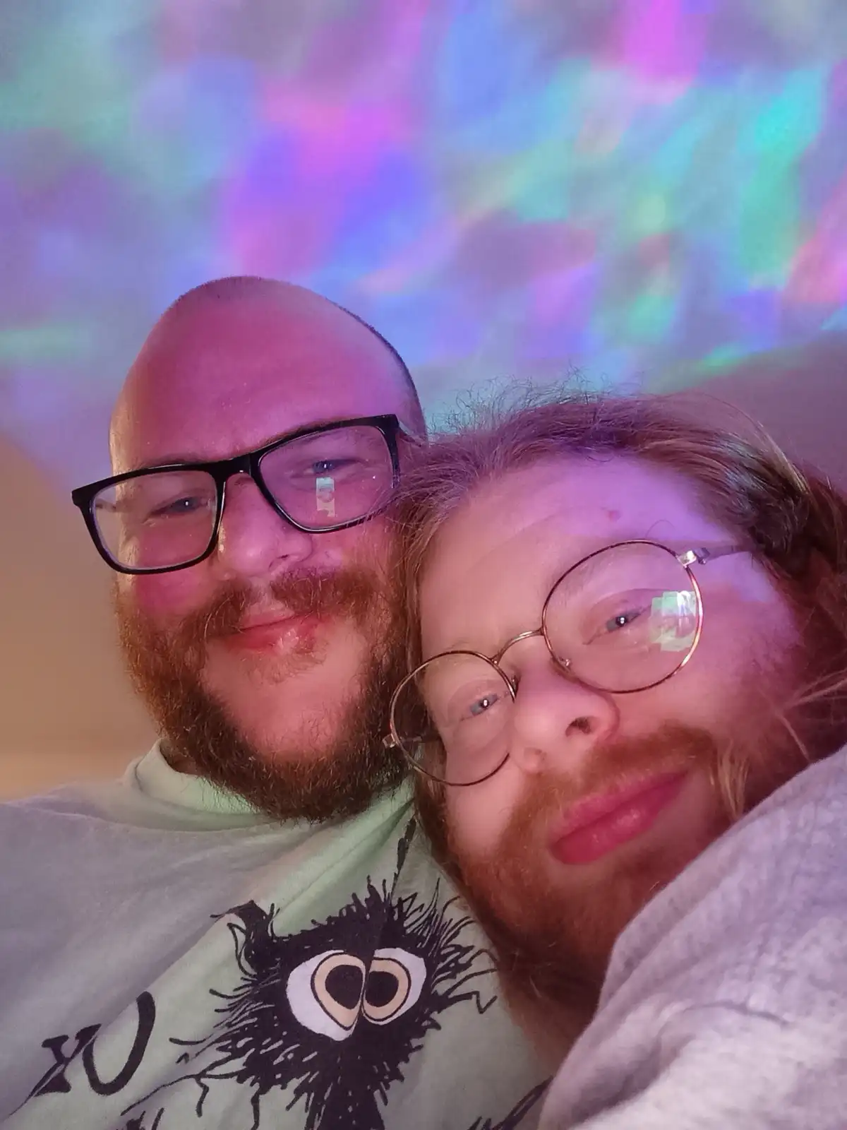 A selfie of two men with reddish beards and glasses, one bald and one with longer hair, smiling closely together under…