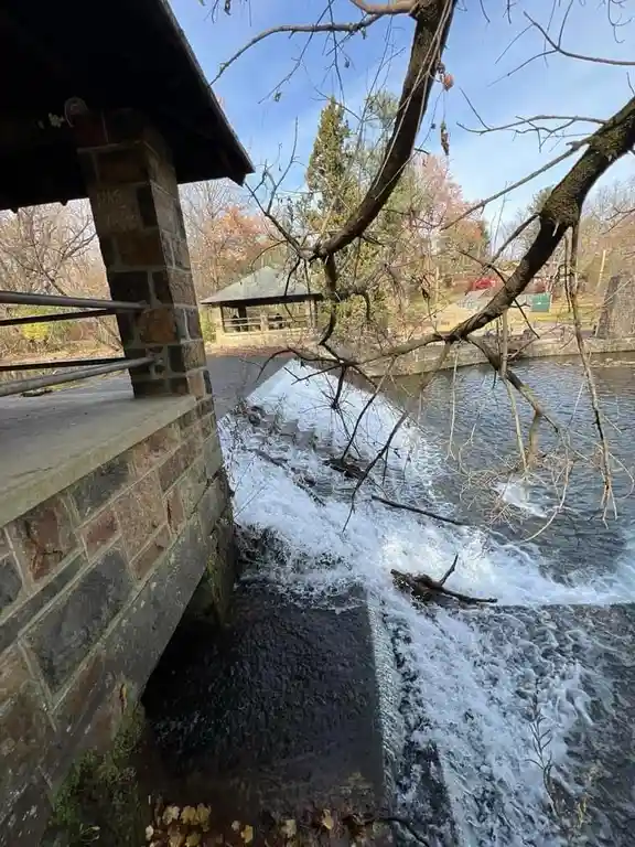 A photo of a small waterfall or weir in a park, with bare tree branches in the foreground and a stone structure on the left…