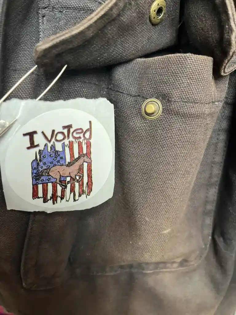 A close-up of a hand-drawn 'I Voted' sticker pinned to a brown jacket. The sticker features a brown horse superimposed over…