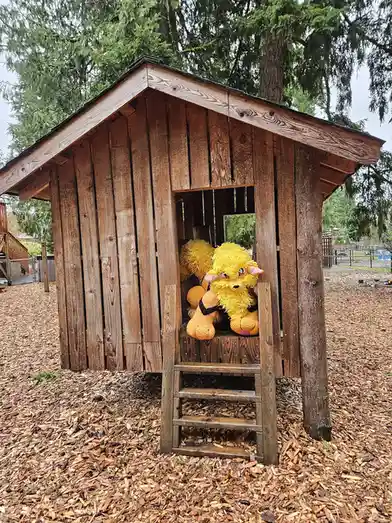 A large, fluffy yellow crocheted stuffed animal resembling a Pokémon sits inside a wooden playhouse with a small ladder…