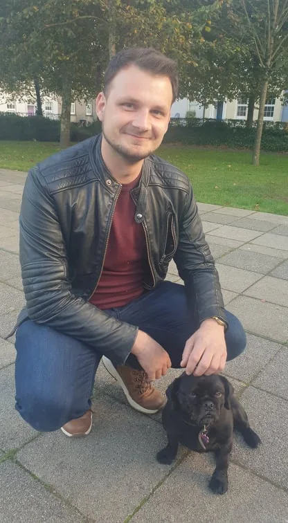 A man in a black leather jacket and jeans is crouching on a paved path outdoors, smiling at the camera while gently petting…