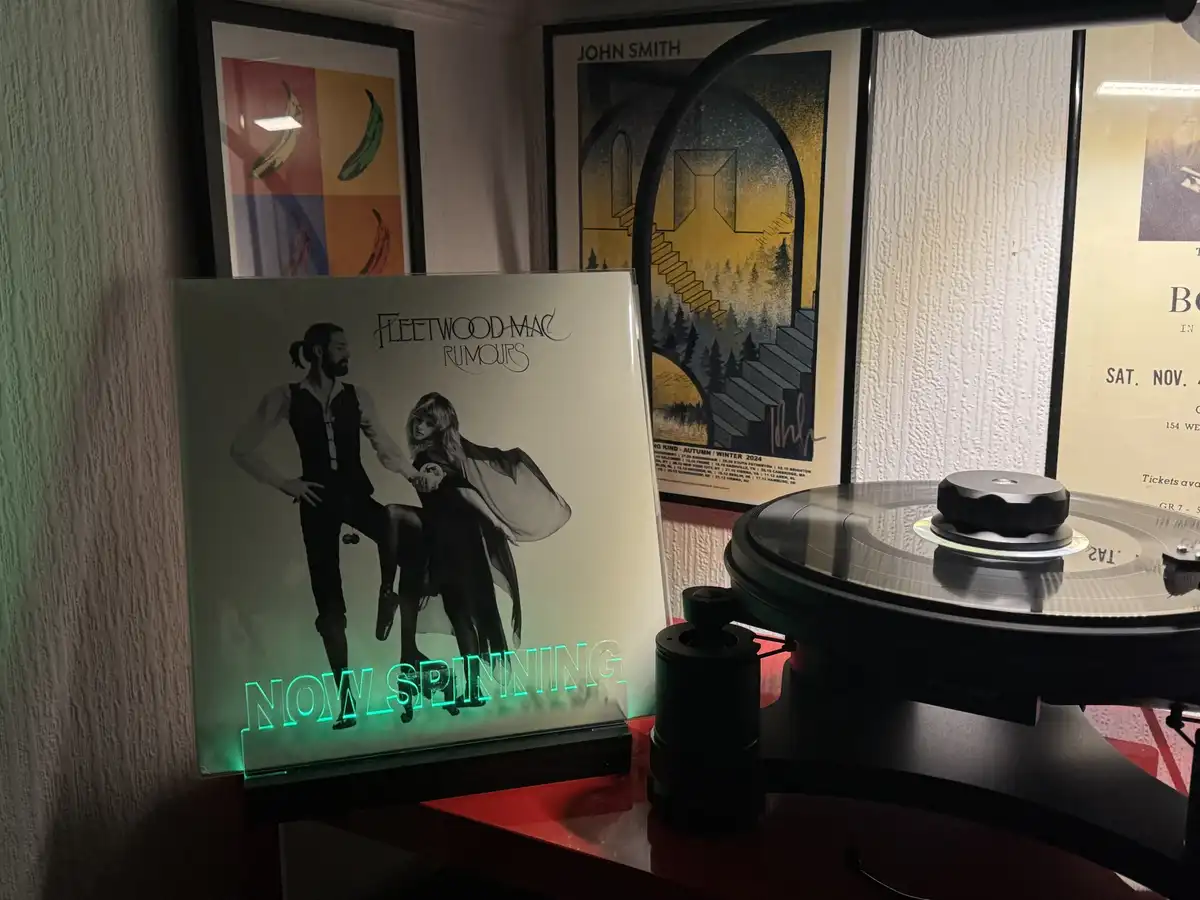 A vinyl record player sits on a red surface next to a glowing neon sign displaying 'NOW SPINNING' beneath a Fleetwood Mac…