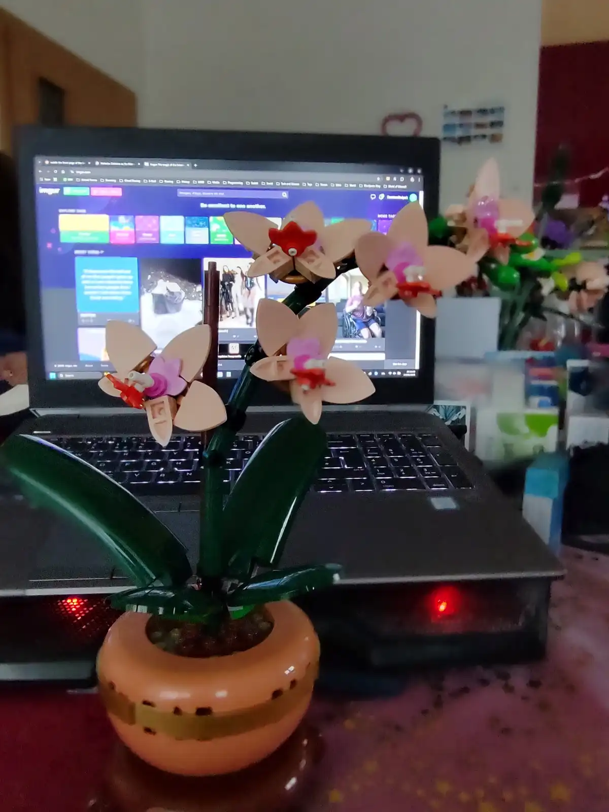 A LEGO-built orchid plant with pink and beige flowers sits in a terracotta pot on a laptop keyboard. The laptop screen…
