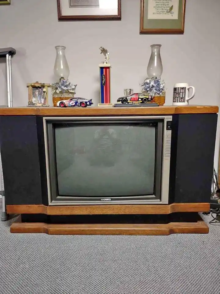 A vintage Magnavox CRT television sits on a wooden console, its screen dark and unlit. The top of the console is cluttered…