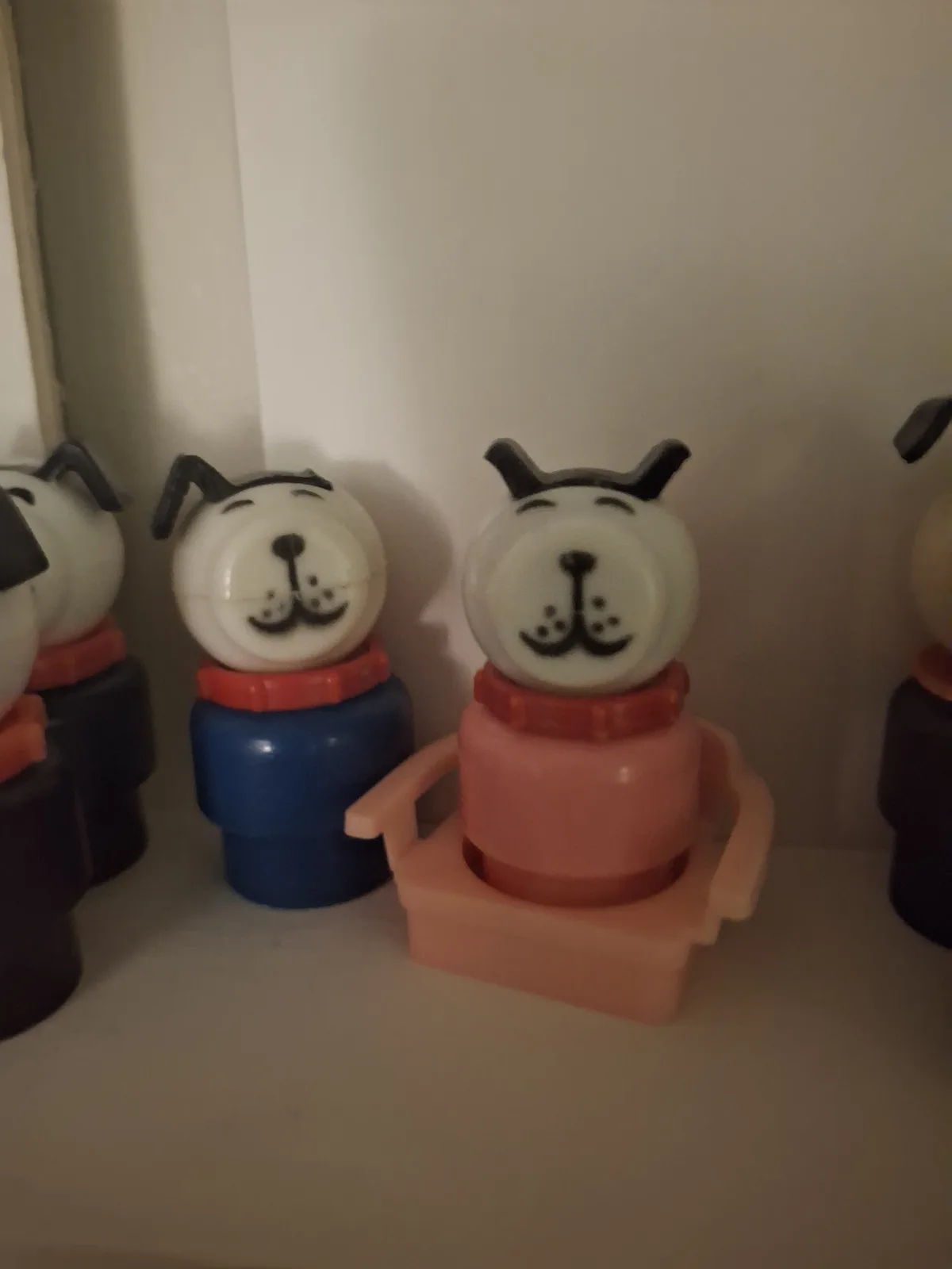 Several plastic dog figurines with simple black facial features and red collars are lined up on a shelf. The central figure…