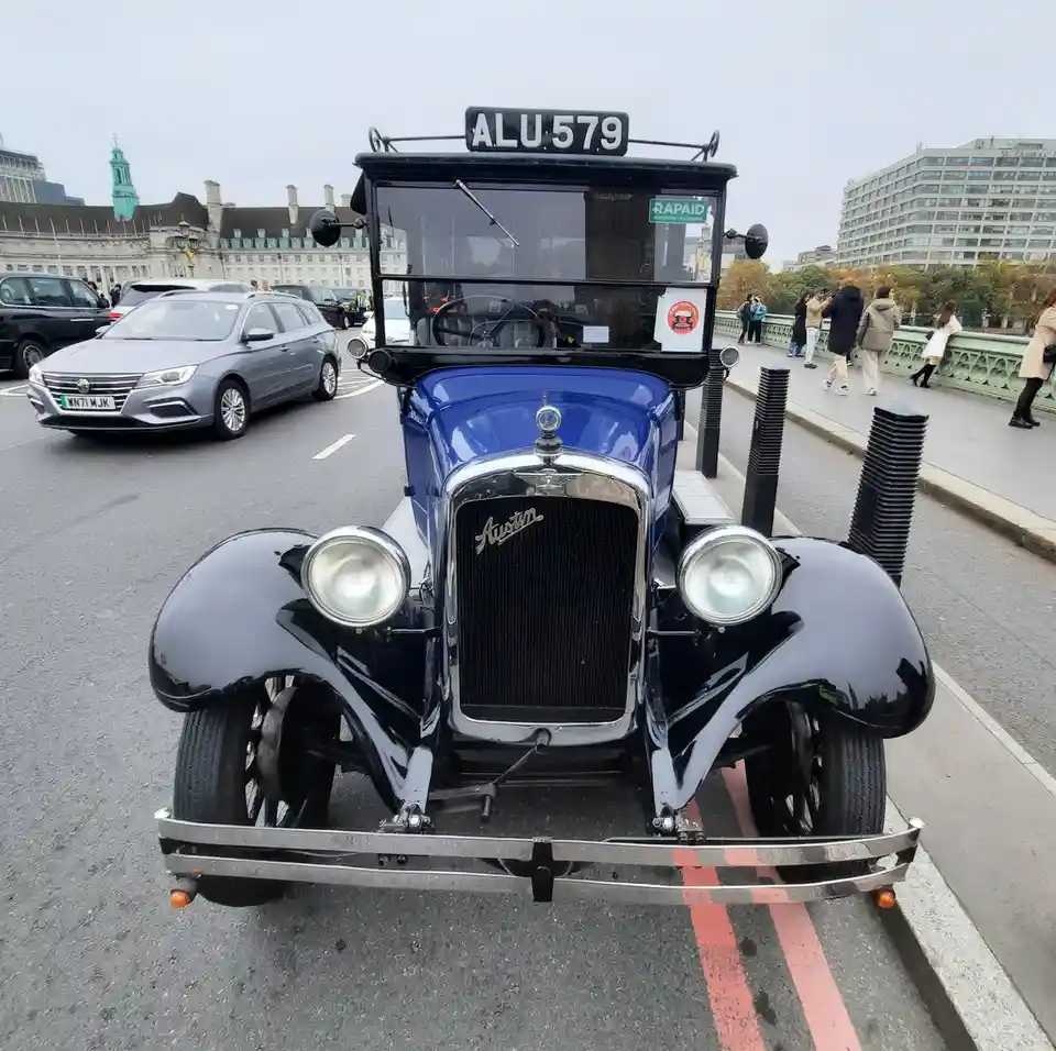 A vintage blue and black Austin car with license plate ALU 579 is driving on a city bridge, with modern cars and pedestrians…