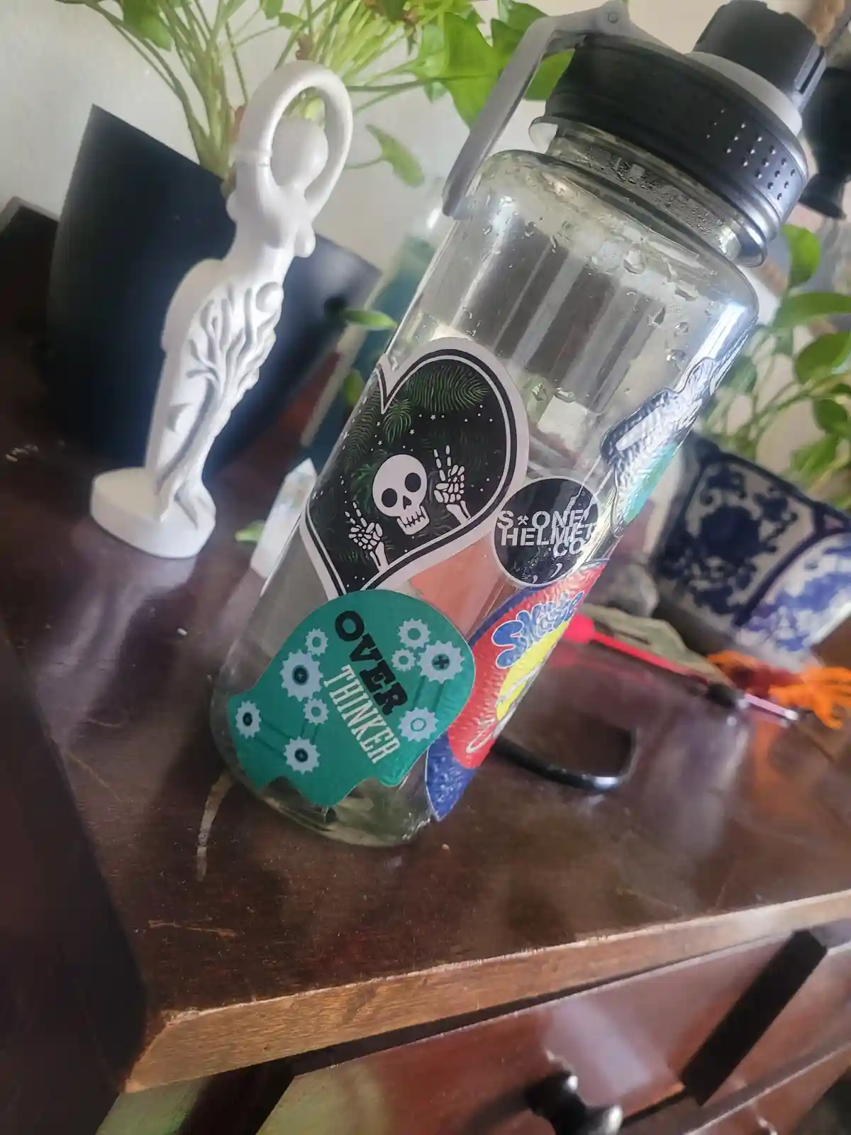 A clear plastic water bottle with a black lid sits on a dark wooden surface, covered in various colorful stickers including…