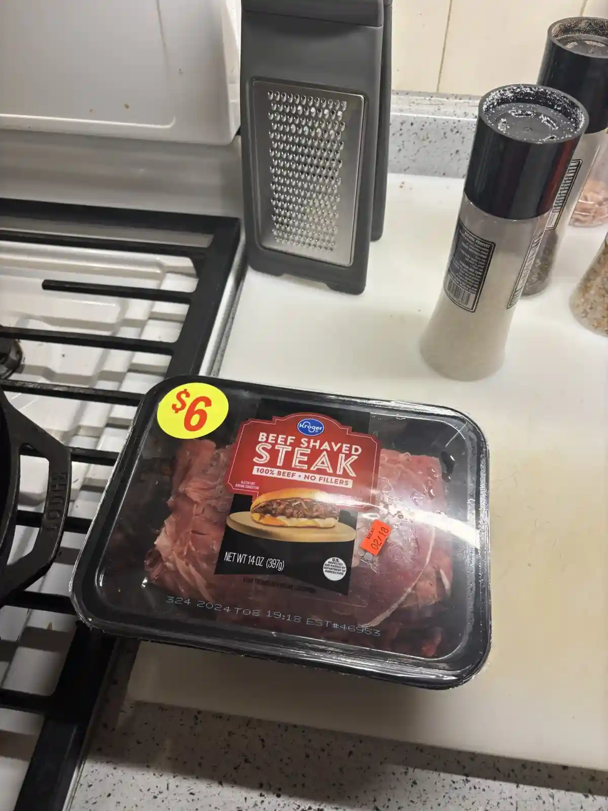 A package of Kroger brand beef shaved steak, priced at $6, sits on a kitchen counter next to a stove, a grater, and salt and…