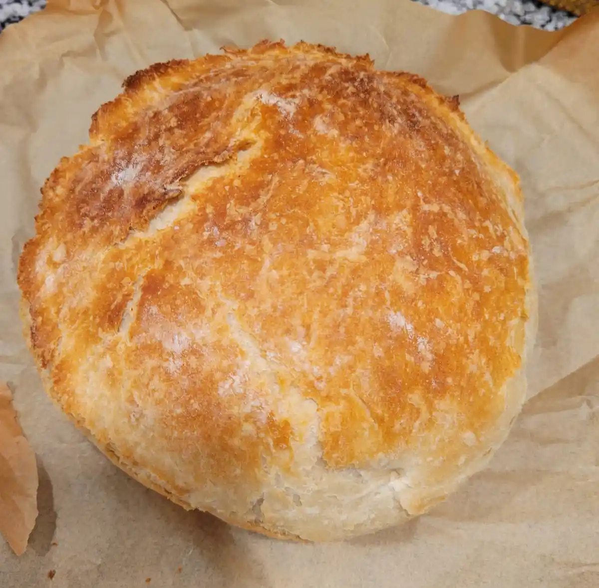 A round, golden-brown loaf of bread with a cracked crust and flour dusting sits on brown parchment paper. The overlaid text…