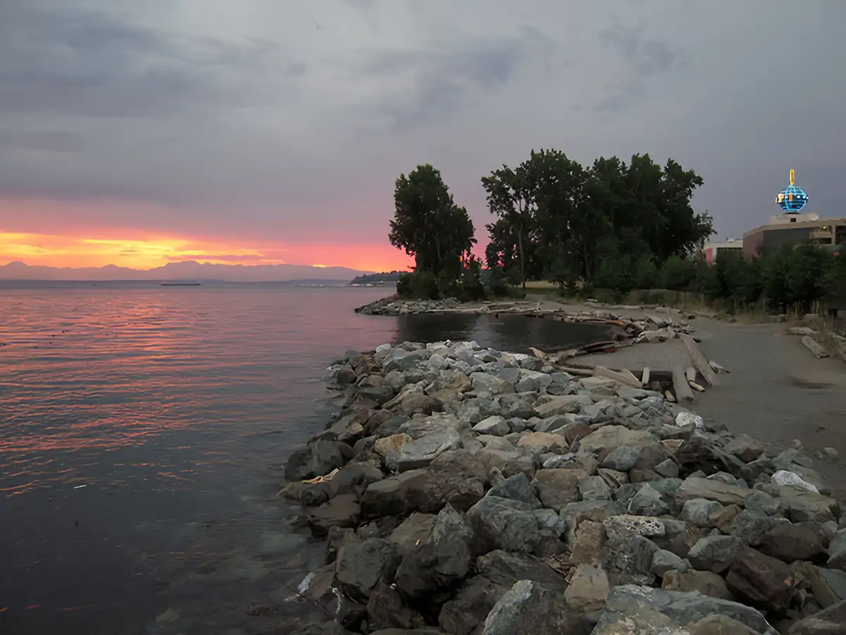 A scenic photo of a rocky shoreline at sunset, with vibrant orange and pink hues reflecting on the water and distant…