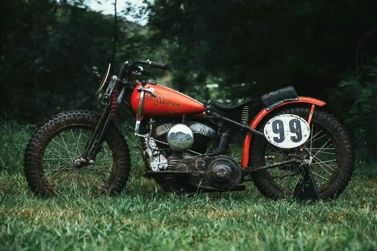 A vintage orange Harley-Davidson motorcycle, covered in mud and grass, is parked on a lawn with trees in the background. The…