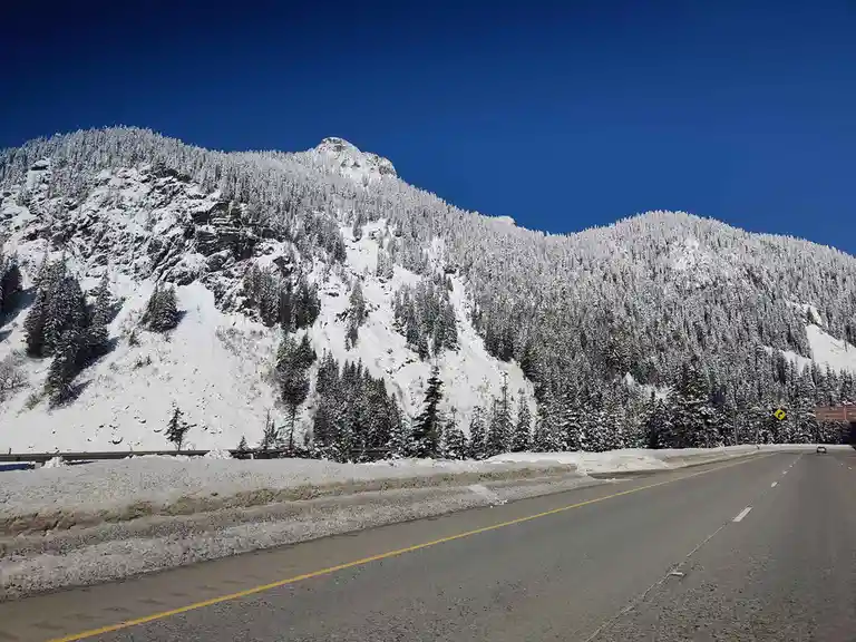 A scenic photo of a snow-covered mountain road under a clear blue sky, with evergreen trees blanketed in snow. A highway…