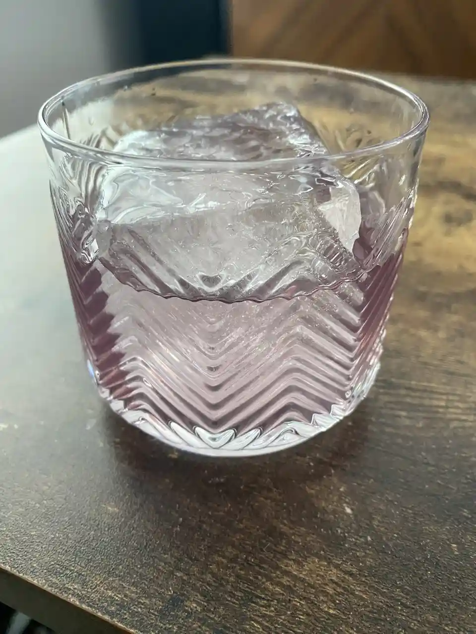A close-up photo shows a clear, patterned glass tumbler sitting on a dark wooden table, containing a single large ice cube…