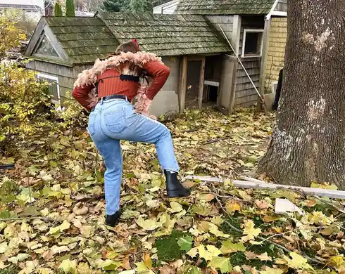 A person wearing a red top, blue jeans, and black boots is seen from behind, bending over in a yard covered in fallen autumn…