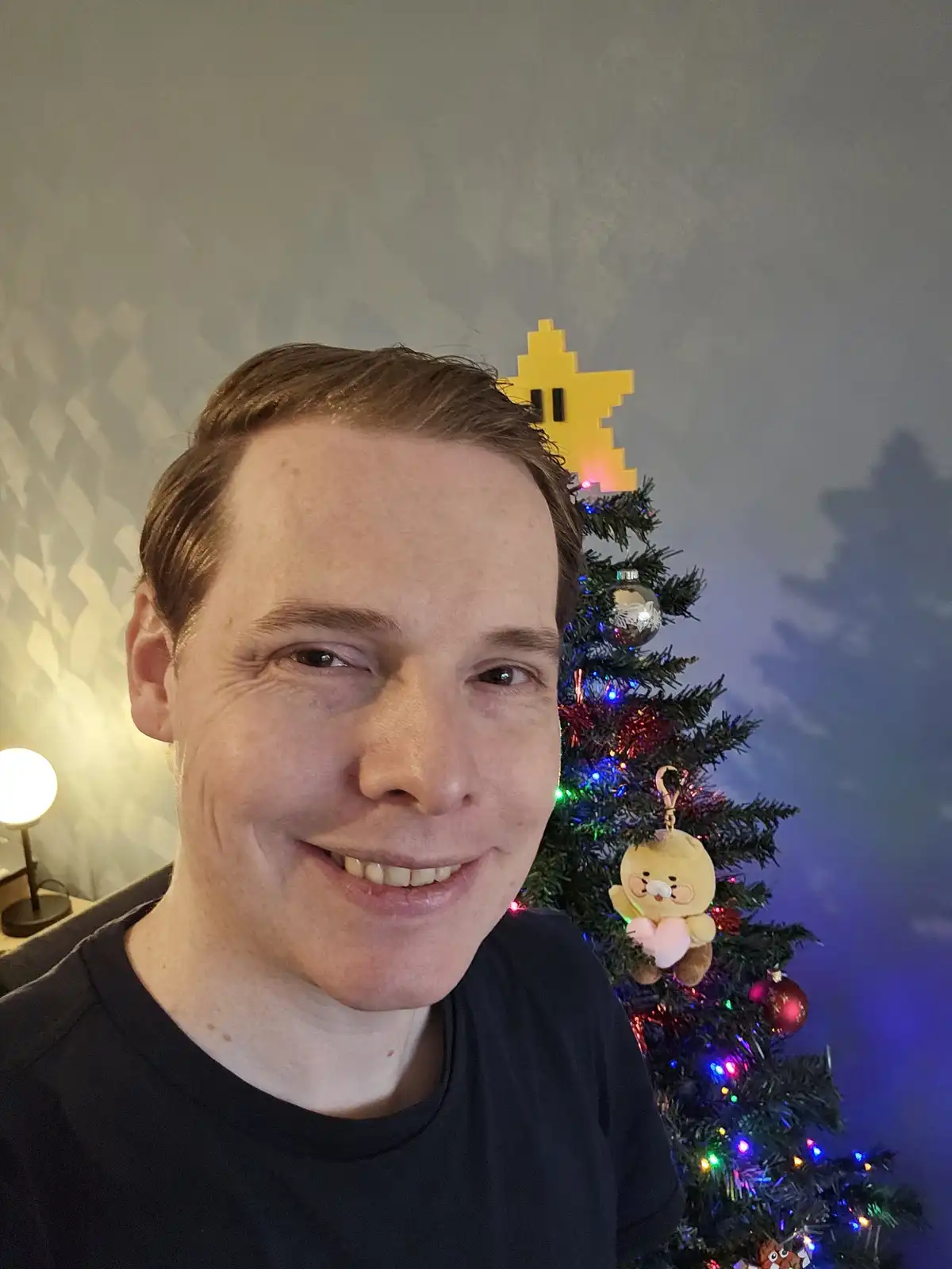 A man smiles for the camera while standing in front of a decorated christmas tree. The tree has colorful lights, ornaments…