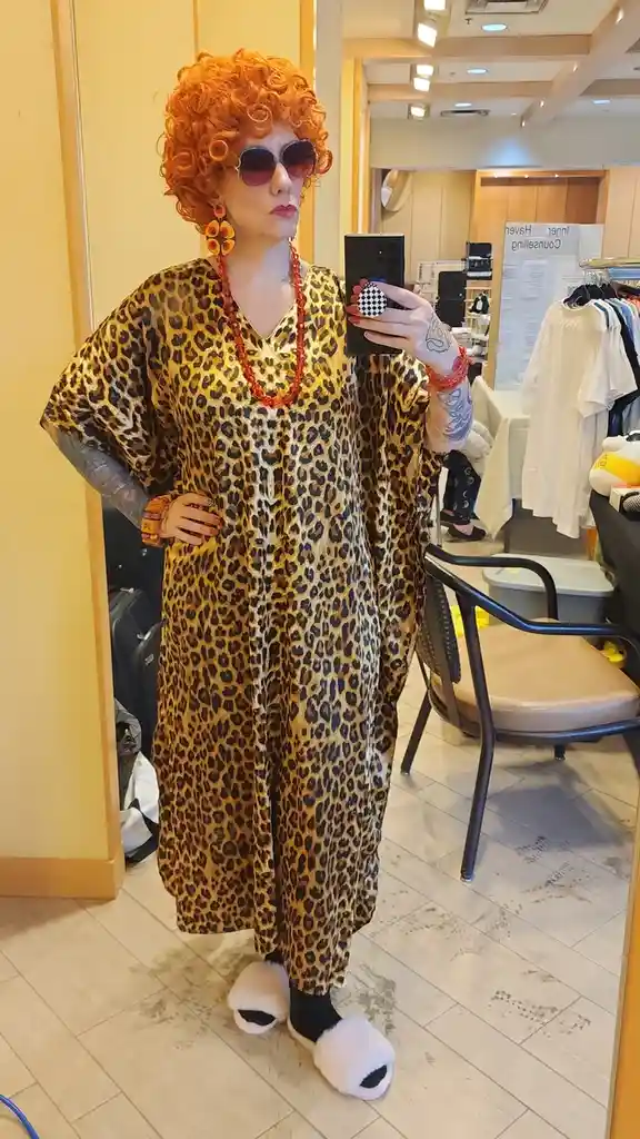 A person with bright orange curly hair and sunglasses takes a mirror selfie while wearing a full leopard-print caftan, red…