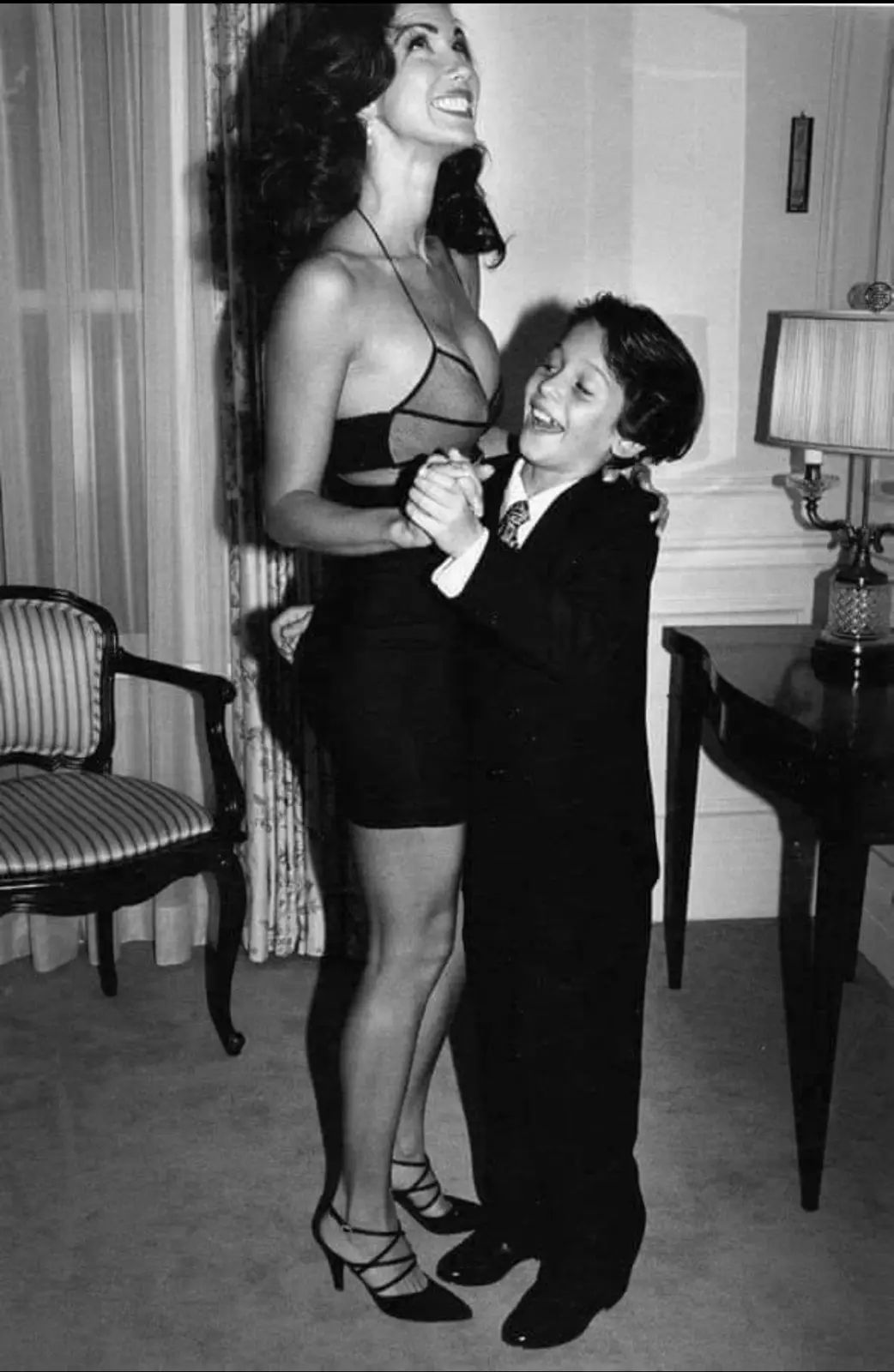 A black-and-white photo shows a tall woman in a revealing dress and heels smiling down at a young boy in a suit who is…