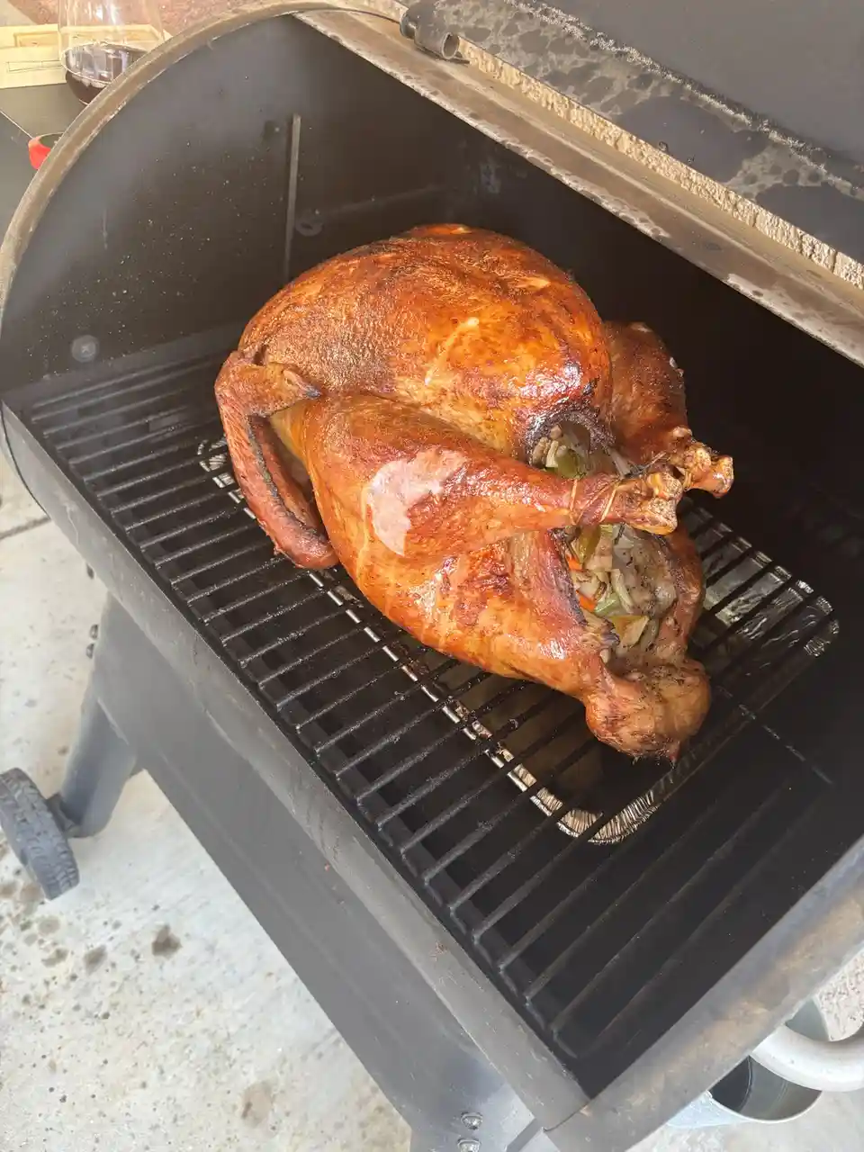 A whole roasted turkey with golden-brown skin sits on the grill grates inside a black smoker or grill. The turkey is stuffed…