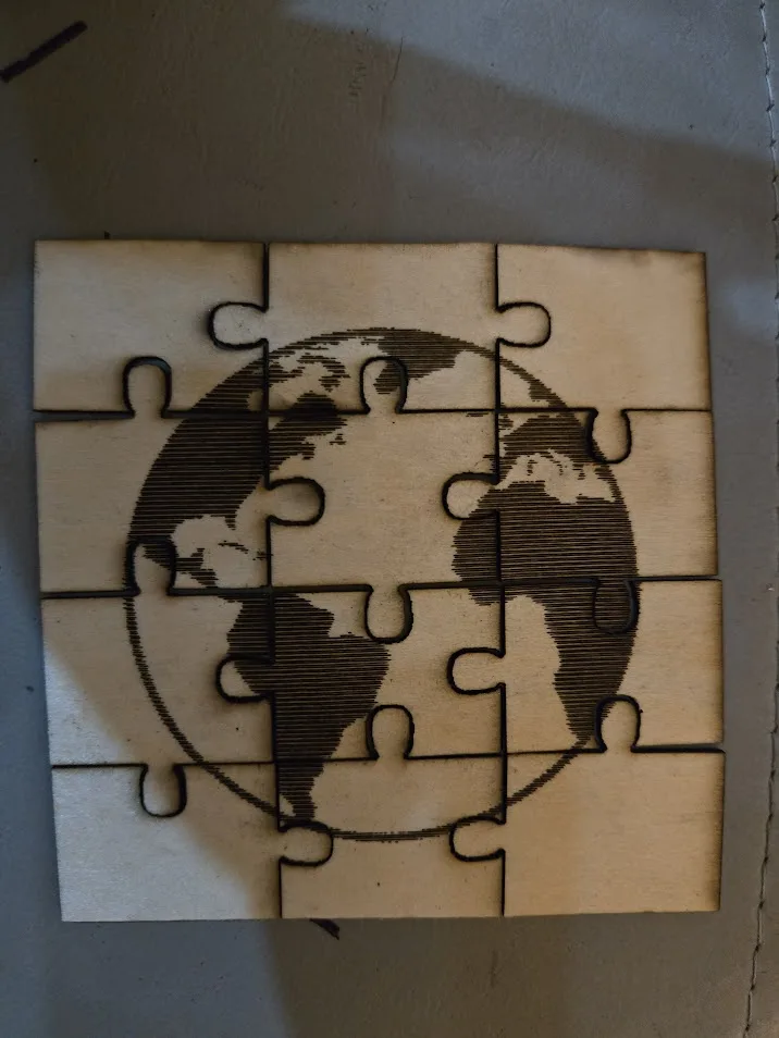 A wooden jigsaw puzzle with a laser-etched world map design, laid out on a gray surface. The text 'the world is a puzzle' is…