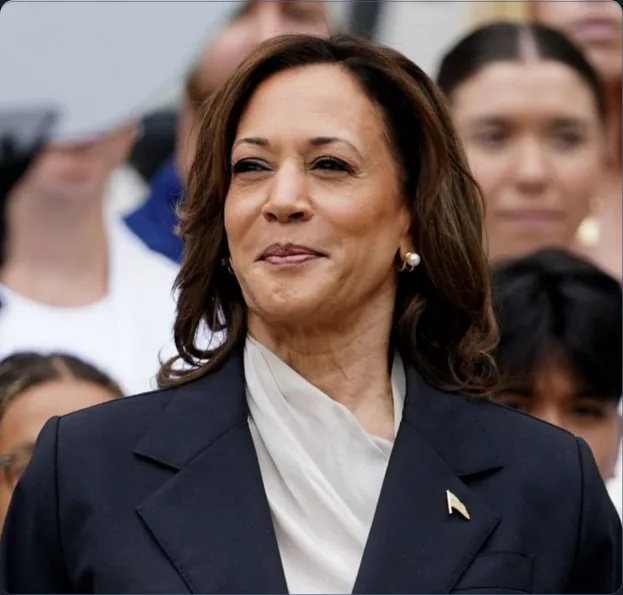 a close-up photo of kamala harris wearing a dark suit and white scarf, looking slightly upward with a subtle smile. The…