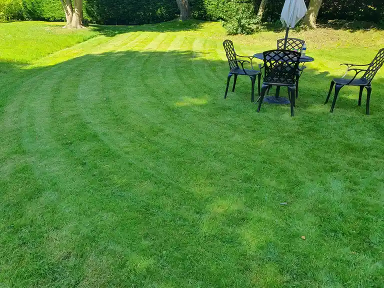 A freshly mowed lawn with visible striped patterns from the mower, featuring a black metal patio set with four chairs and a…