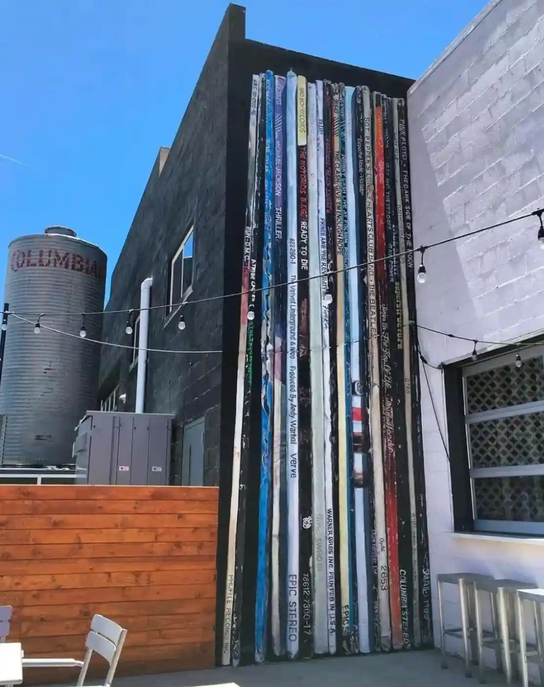 An outdoor patio area features a wall constructed from vertically stacked, weathered vinyl record sleeves, displaying album…