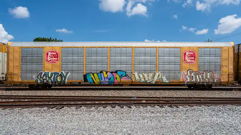 A yellow Kansas City Southern Lines freight train car sits on tracks under a blue sky with clouds, covered in colorful…