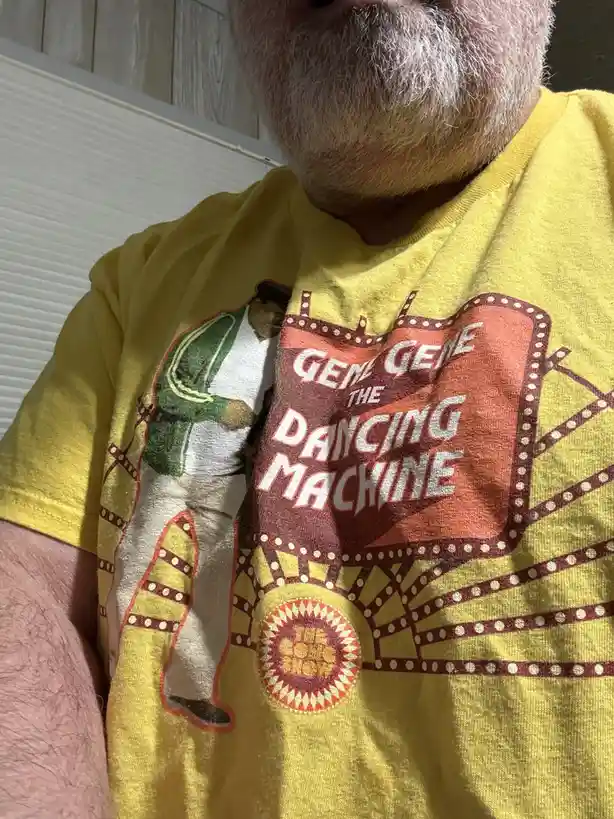 A close-up photo shows a man with a white beard wearing a yellow t-shirt featuring a vintage-style graphic of Gene Kelly in…