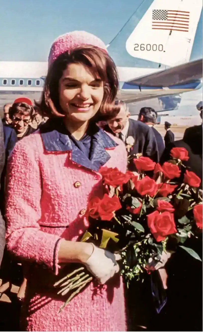 a woman in a pink tweed suit and matching hat smiles while holding a large bouquet of red roses, standing in front of an…