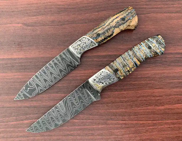 Two intricately crafted knives with damascus steel blades and ornate, patterned handles rest diagonally on a reddish-brown…
