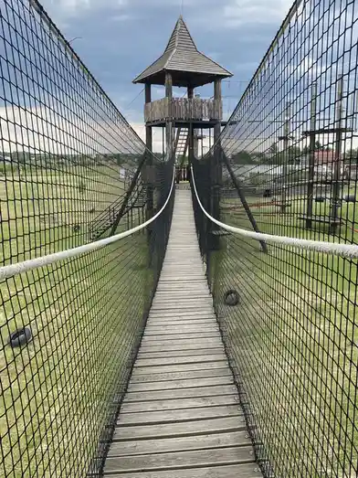 A wooden suspension bridge with black safety netting on both sides leads to a wooden tower with a pointed roof. The bridge…