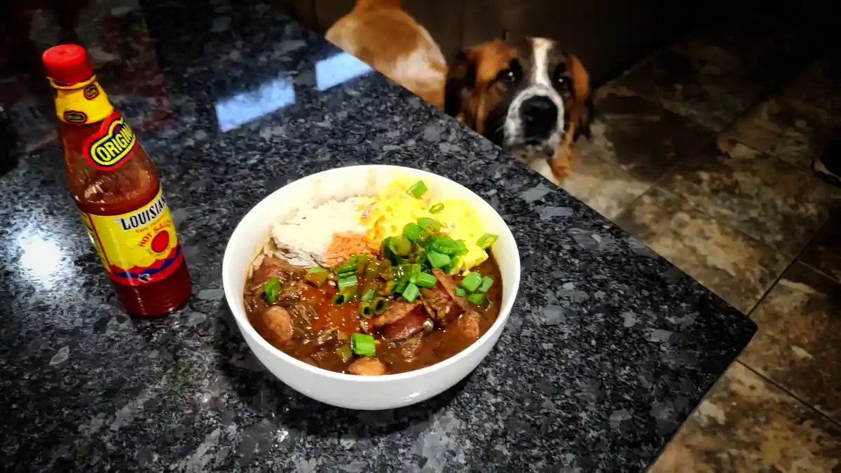 A large bowl of jambalaya with rice, scrambled eggs, and green onions sits on a dark granite countertop next to a bottle of…