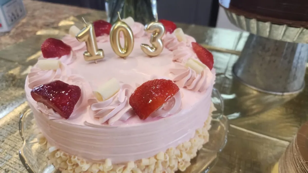A close-up of a pink-frosted birthday cake decorated with piped frosting, fresh strawberries, and white chocolate curls…