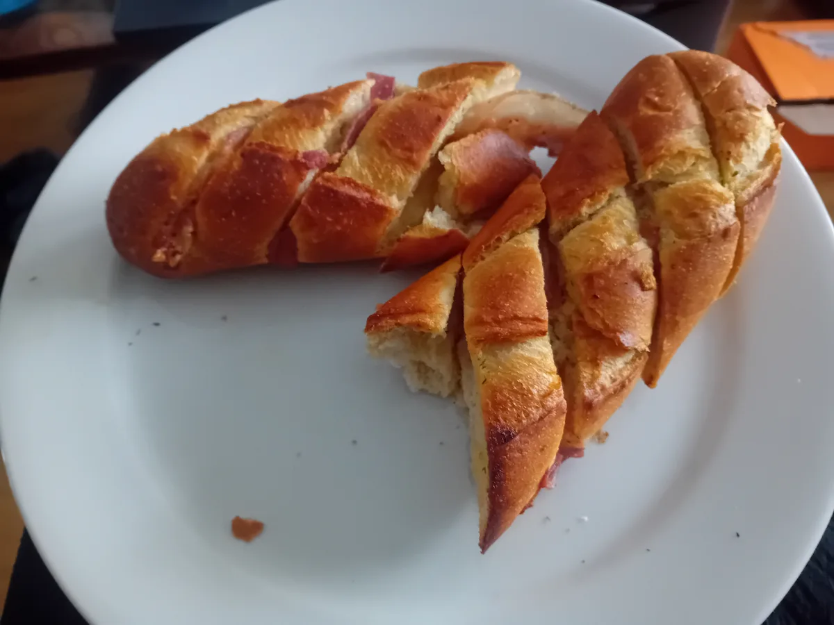 A partially eaten braided bread roll with ham filling sits on a white plate, with a few slices removed and crumbs scattered…