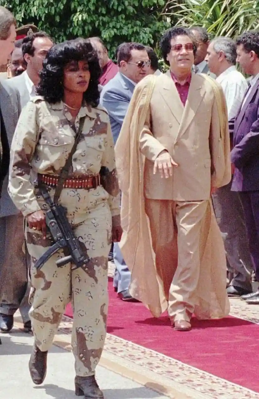 A historical photograph shows Muammar Gaddafi in a tan suit walking beside his wife, Sa'ra Gaddafi, who is in military…
