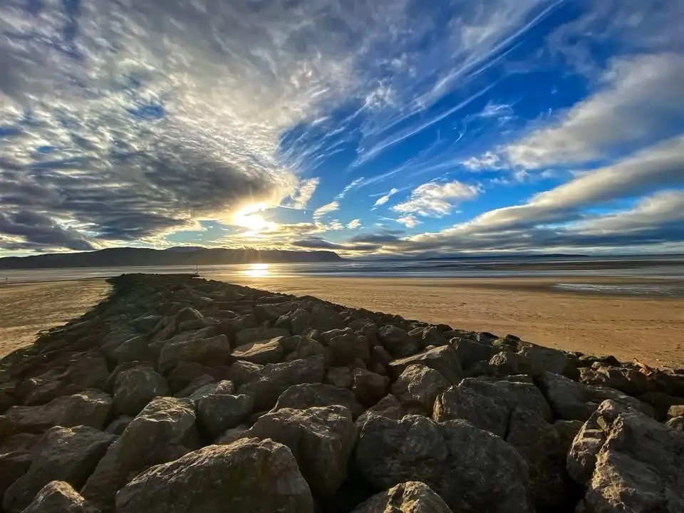 A scenic photo of a rocky breakwater leading into a wide sandy beach at sunset, with dramatic clouds and golden light…