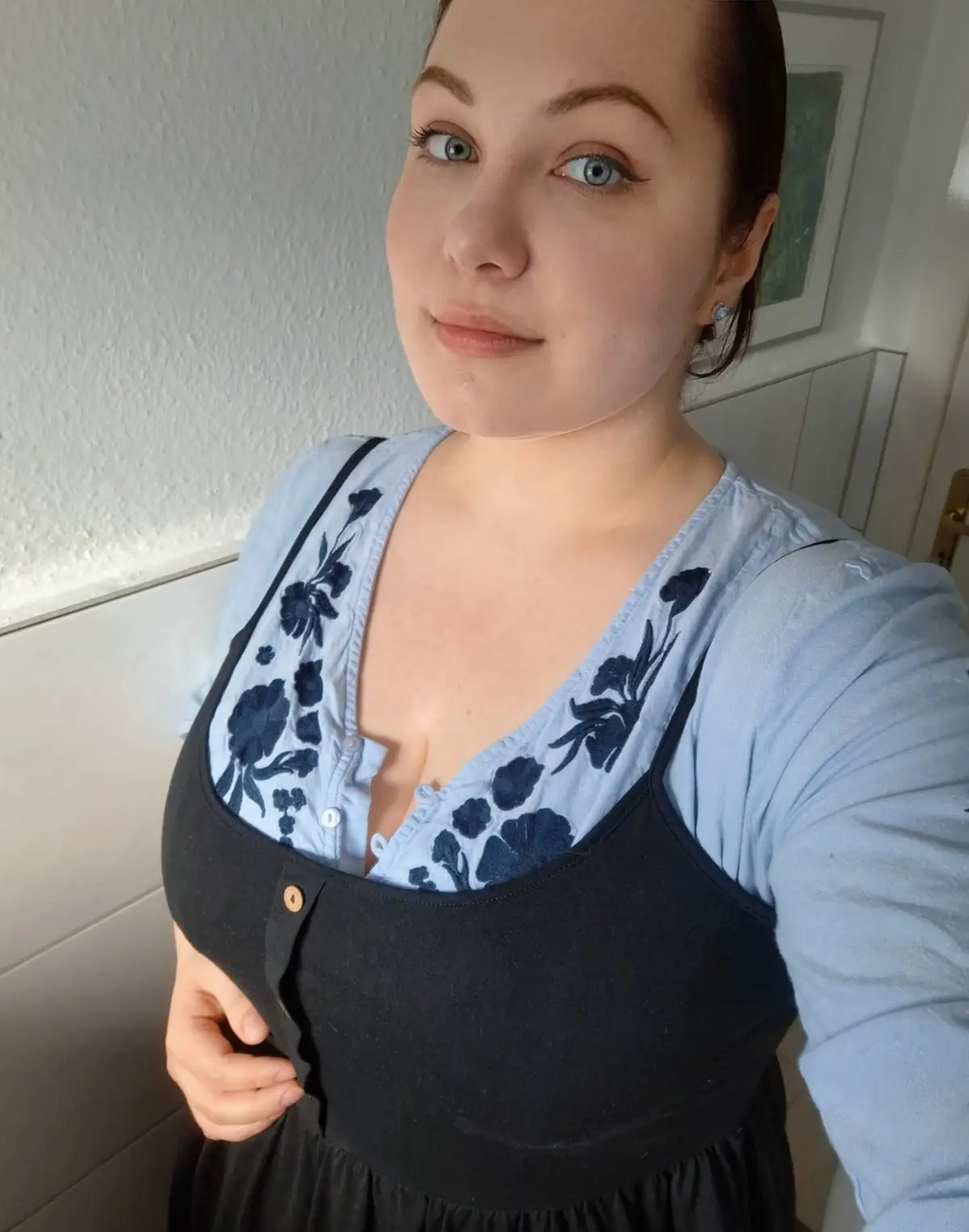 A woman with blue eyes and dark hair pulled back looks at the camera with a slight smirk while wearing a light blue floral…