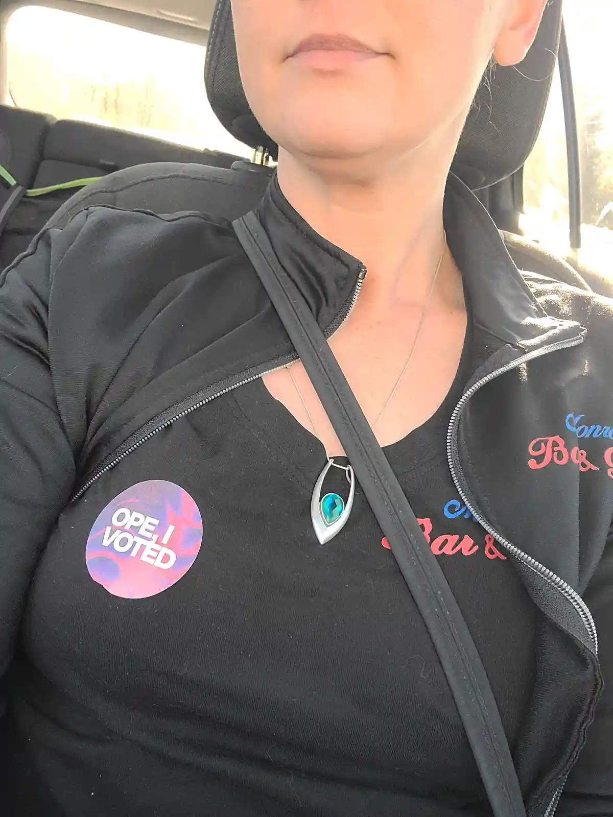 A person sitting in a car wears a black zip-up jacket over a black shirt, with a circular 'OPE, I VOTED' sticker on their…