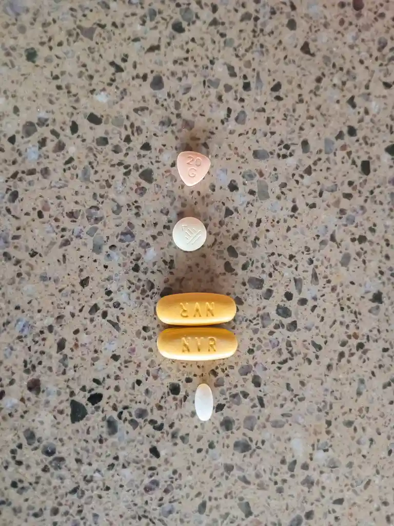 Five pills of varying shapes, sizes, and colors are arranged vertically on a speckled countertop. From top to bottom: a…