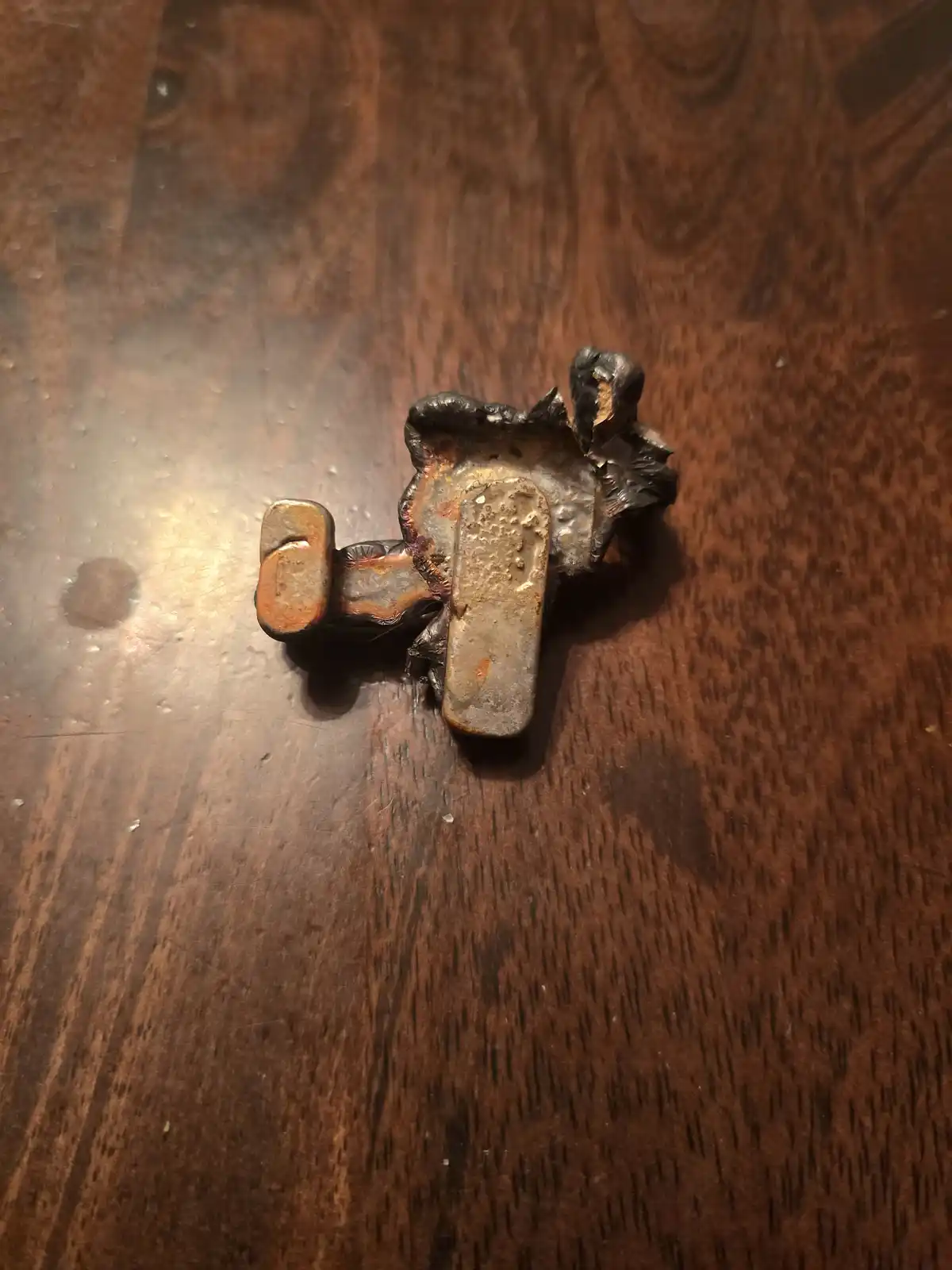 A small, melted metal figurine, possibly a toy or pin, lies on its back on a dark wooden surface, showing distorted, melted…