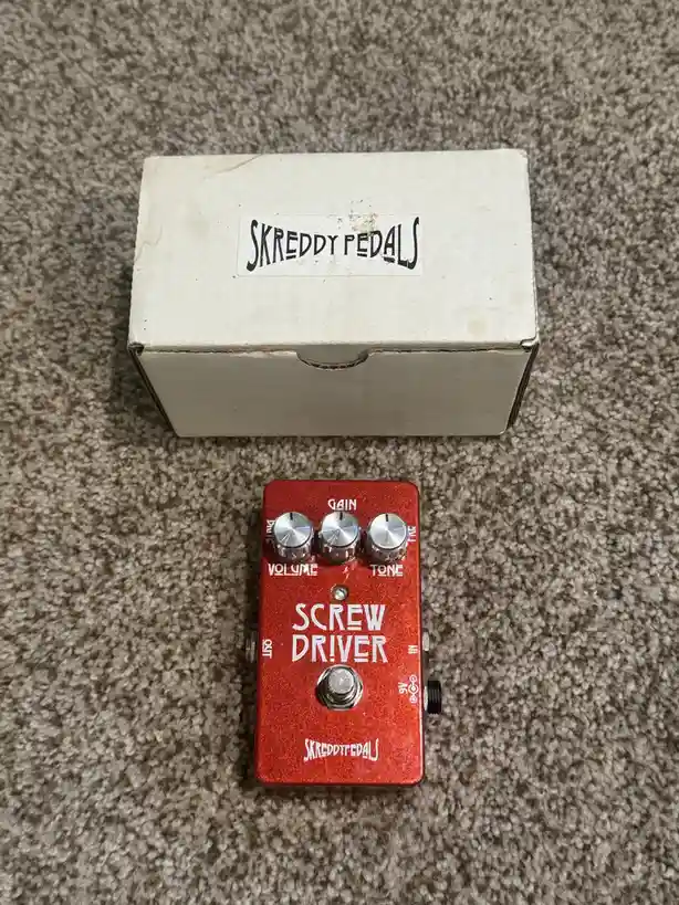 A red guitar effects pedal labeled 'SCREW DRIVER' by 'SKREDDY PEDALS' sits on carpet next to its white box, which also has…