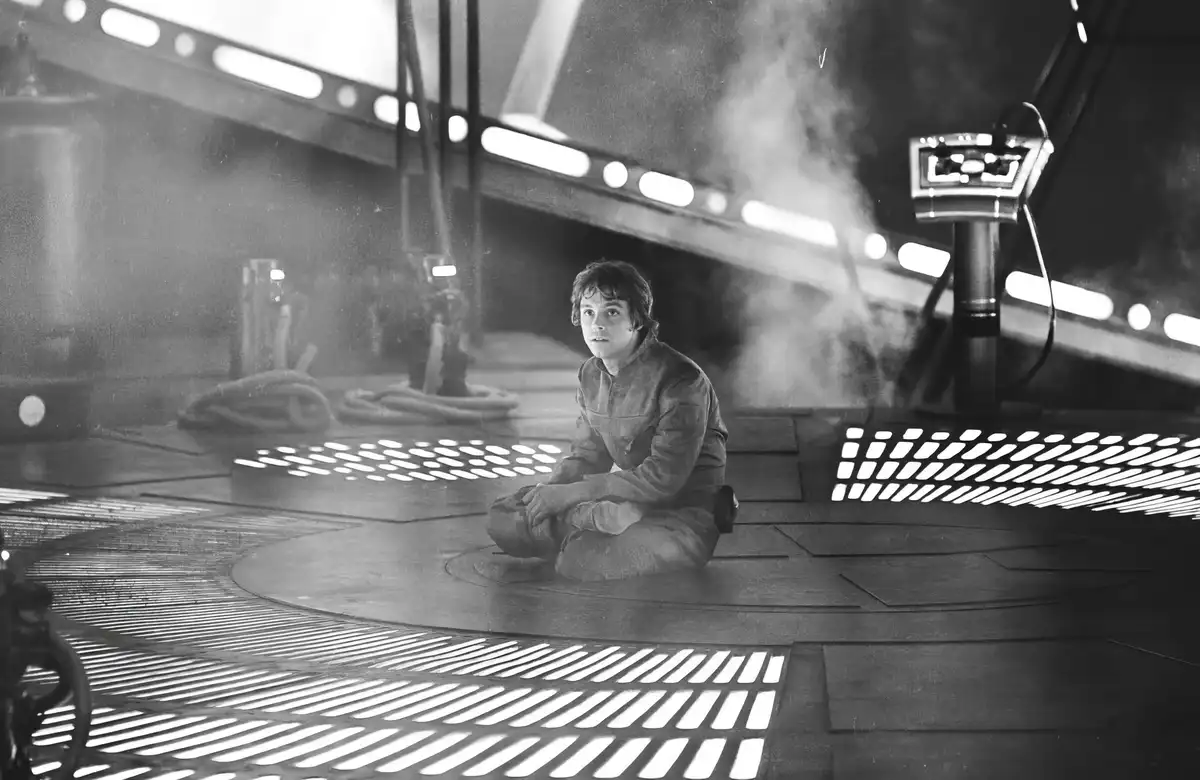 A black-and-white image shows a young man sitting cross-legged on a grated floor in a futuristic, industrial-looking room…