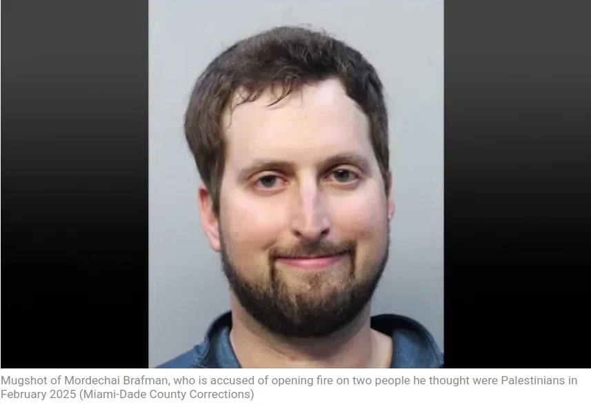 A mugshot of a man with short brown hair and a beard, wearing a blue shirt, smiling slightly at the camera. The image has a…