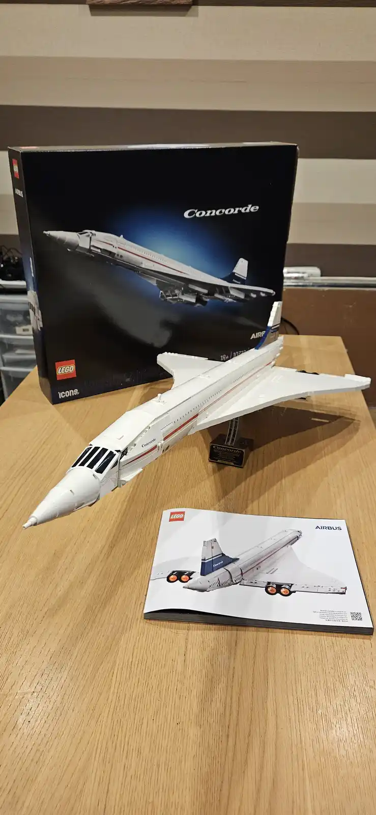 A completed white LEGO model of the Concorde supersonic jet airplane sits on a wooden table, displayed next to its black box…