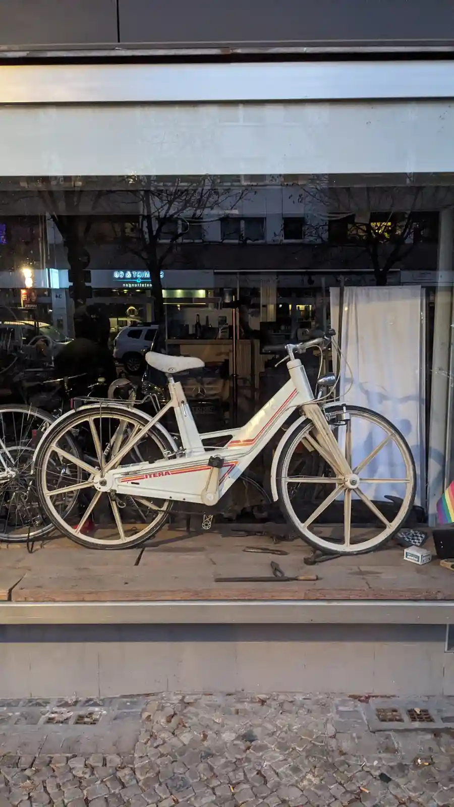 A white vintage bicycle with orange stripes and the brand name 'ITERA' is displayed in a shop window, surrounded by tools…