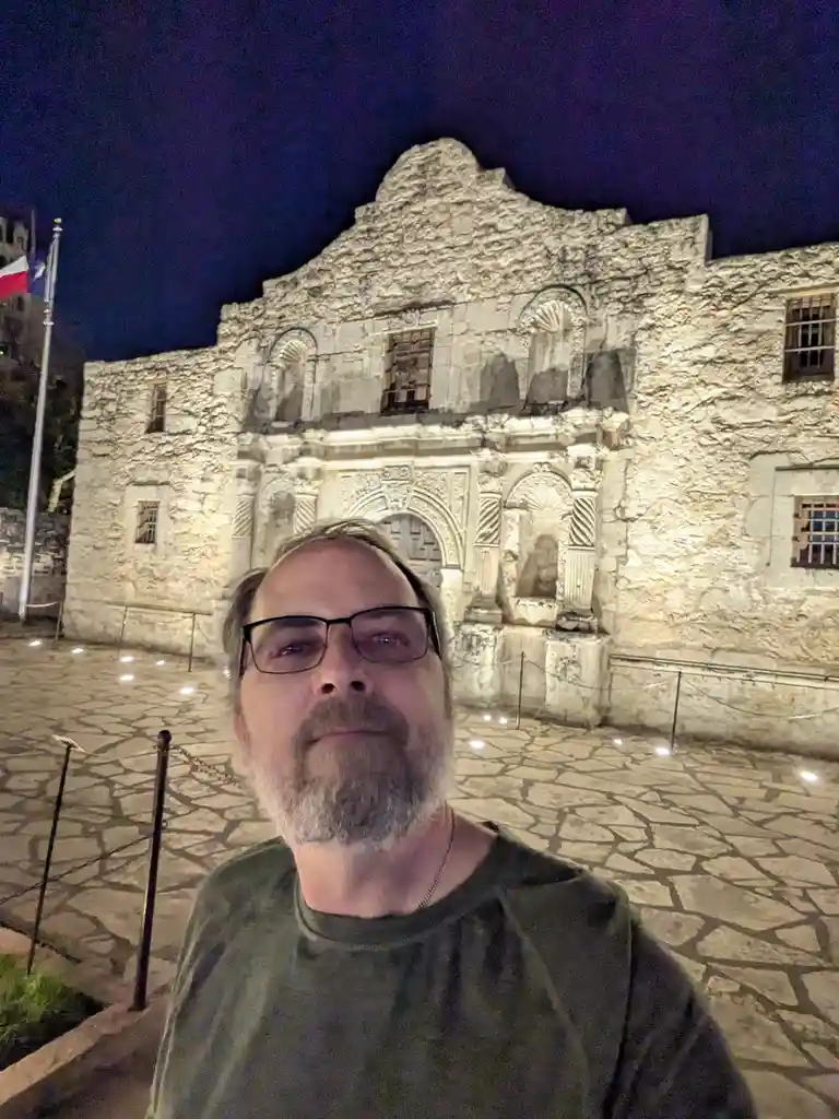 A man with glasses and a beard takes a selfie at night in front of the historic Alamo in San Antonio, Texas. The stone…