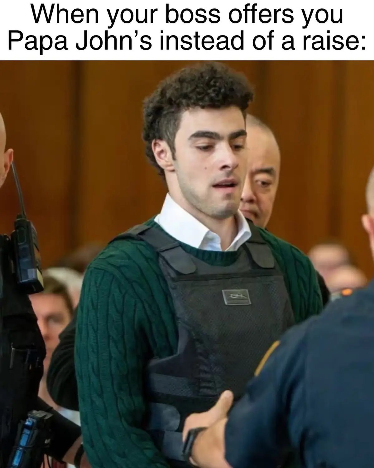 A man with curly dark hair, wearing a green cable-knit sweater under a black tactical vest, looks downcast and disappointed…