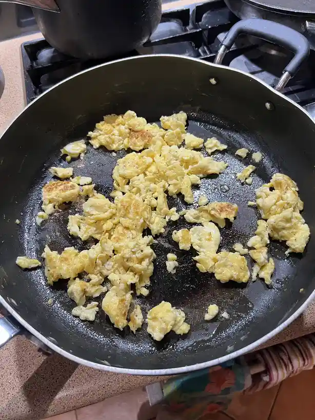 A close-up photo shows scrambled eggs in a black frying pan on a stovetop, with the eggs appearing lumpy and unevenly…