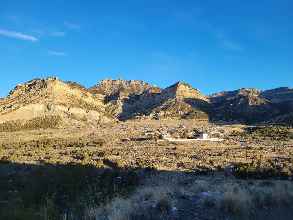 A scenic photo of a small town nestled in a valley surrounded by large, sunlit mountains under a clear blue sky. The…