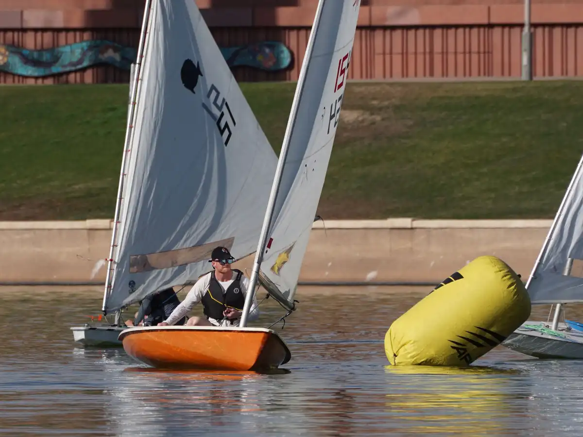A man in a small orange sailboat wearing a life vest and sunglasses looks toward a large yellow inflatable buoy partially…